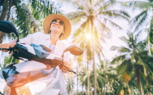 Happy smiling woman in a straw hat and sunglasses riding motorbike under palm trees